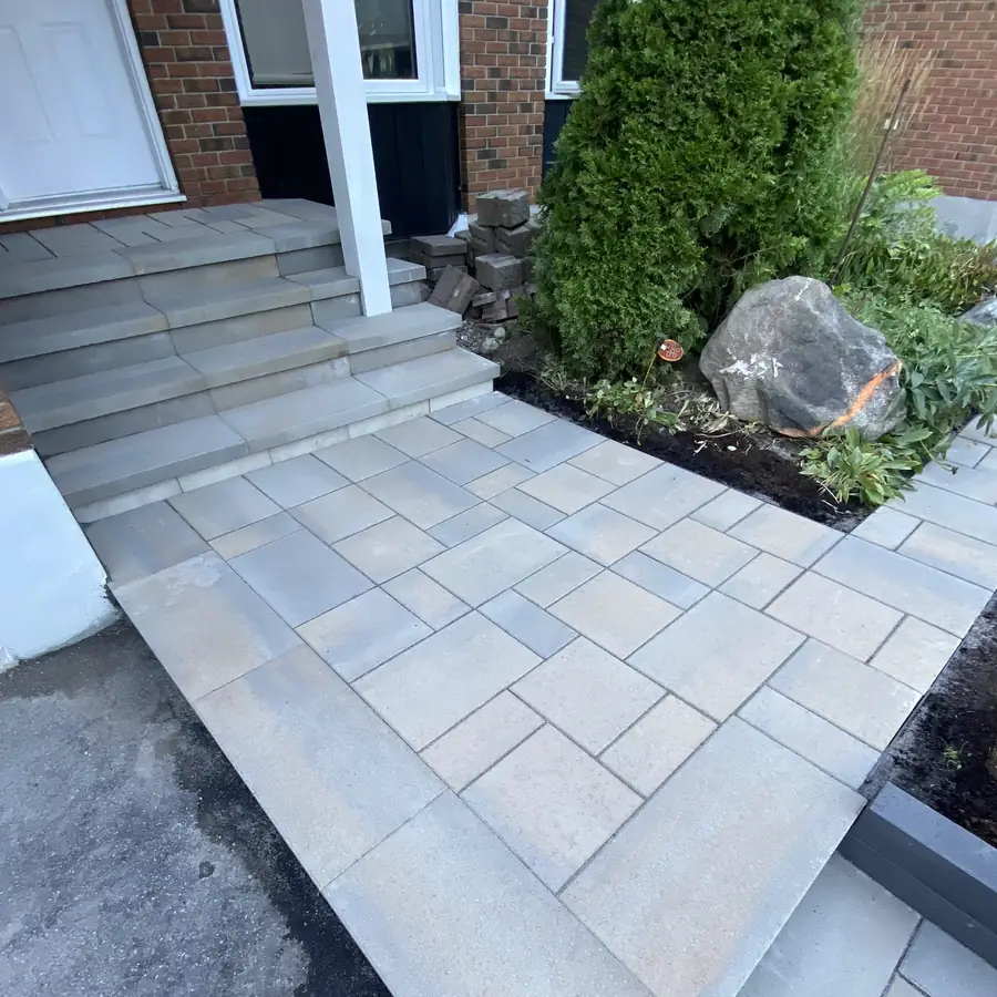 A new grey interlock walkway and steps leading to a front door, with a stone border and plants beside it