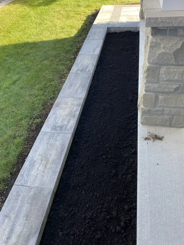 A raised garden bed of dark soil edged in pale grey stone, beside a stone pillar