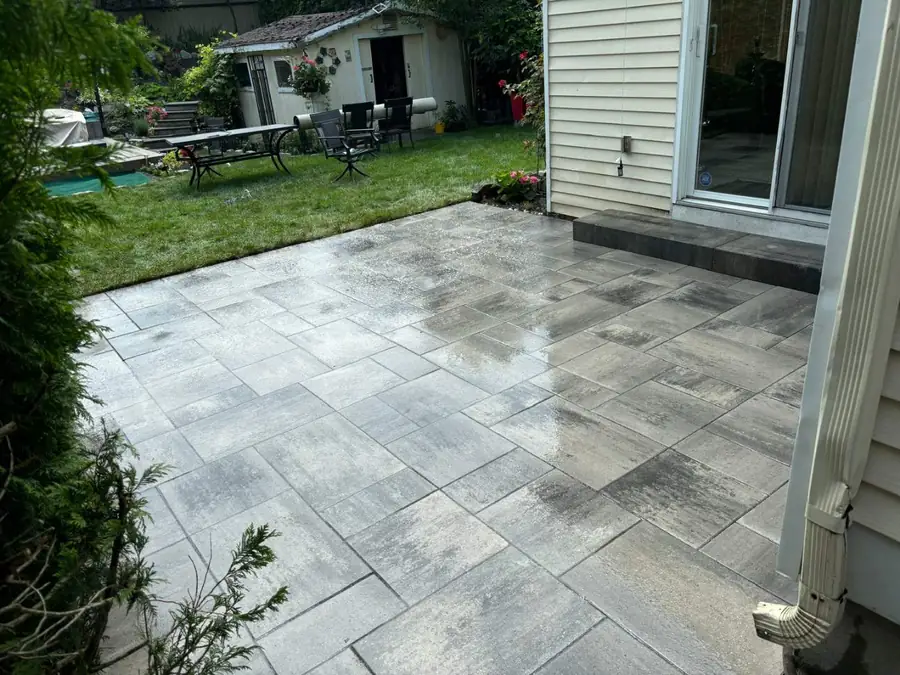 A backyard patio of large grey pavers, darkened by rain, beside the house